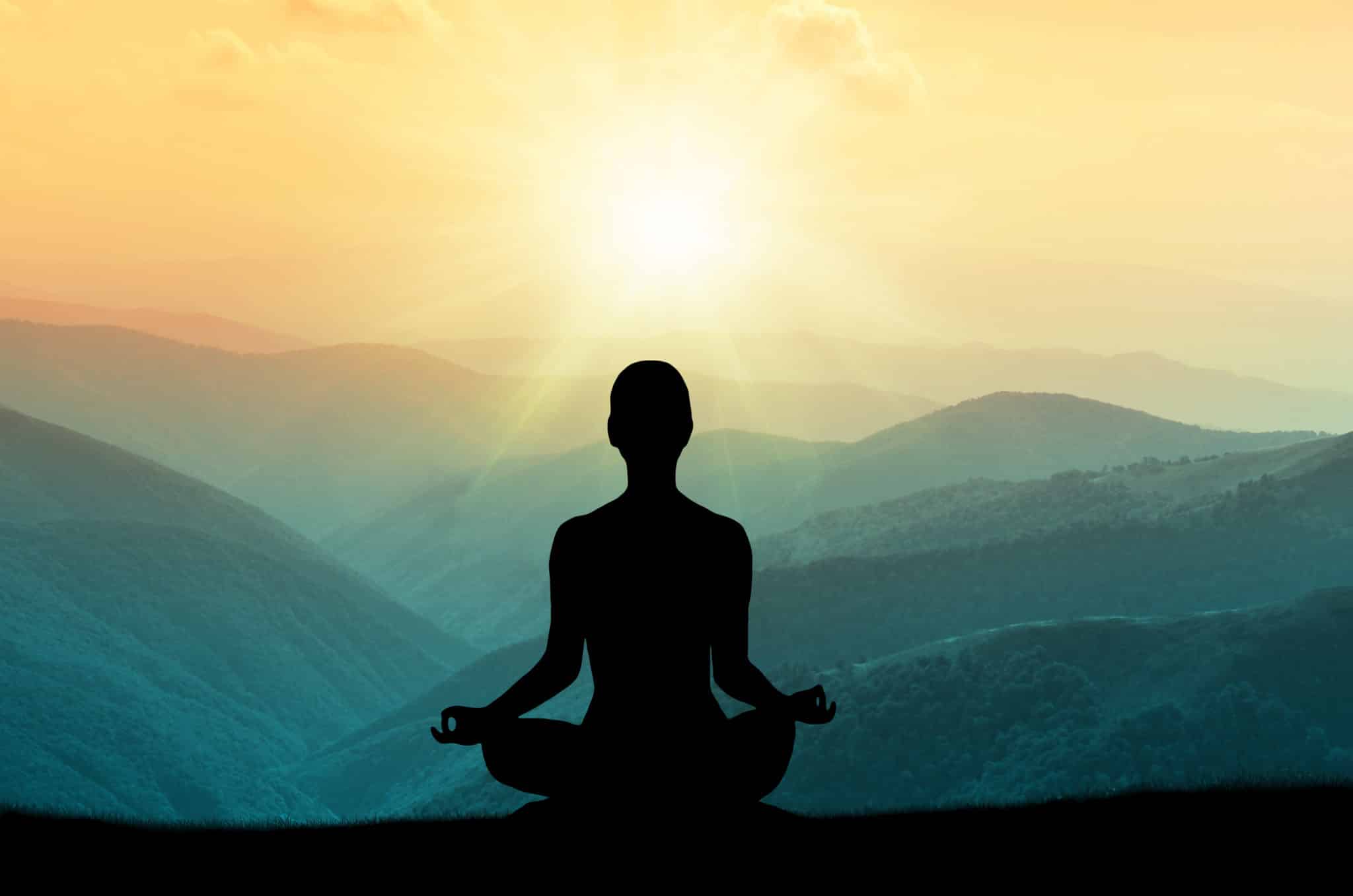 Woman sitting in meditation pose outdoors looking at the mountains and sunset